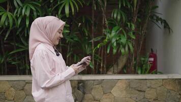 moderno musulmano donna su hijab sud-est asiatico - a passeggio, sorridente, guardare sua smartphone all'aperto video