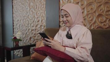 modern moslim vrouw Aan hijab zuidoosten Aziatisch - lachend, op zoek en scrollen haar smartphone terwijl zittend Aan bankstel in de leven kamer video