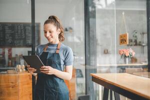 Beautiful young barista woman in apron holding tabltet and standing in front of the door of cafe with open sign board. Business owner startup concept. photo