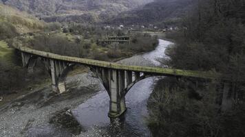 Aerial of abandoned concrete bridge covered by moss over the narrow river. Shot. Autumn landscape with high forested mountains and the small village. photo