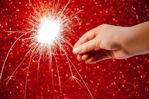 New Year celebration. Womans hand holds bright sparkler on red background. Christmas night. Bright sparks of fire and bokeh. photo