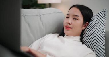 Footage of Happy young Asian woman using a laptop computer while lying on the sofa in the living room. Wellness at home, relaxing and lifestyle concepts. video