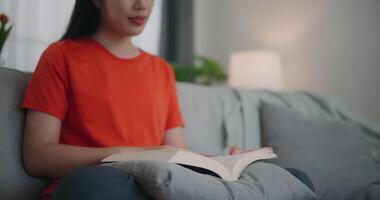 imágenes de contento joven asiático mujer leyendo un libro mientras sentado en el sofá en el vivo habitación. bienestar a hogar, relajante y estilo de vida conceptos. video