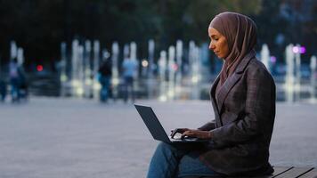 Young successful muslim business woman islamic student girl wears hijab sitting on street city outdoors urban background typing laptop working online e-learning outside with computer chatting app web photo