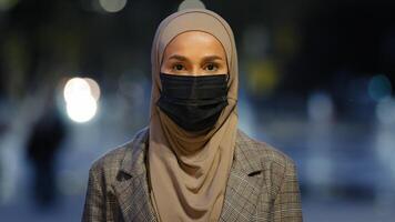 Close-up islamic female in medical mask posing in night city. Muslim business woman wearing hijab masked girl standing looking at camera evening urban background outdoors pandemic coronavirus covid photo