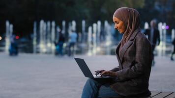 Busy successful muslim student girl in hijab islamic business woman freelancer using laptop app working in city fountains background typing email in net. Ethnic female with wireless computer study photo