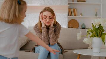 Tired upset Caucasian mother sitting on sofa at home sad frustrated stressed exhausted fatigued mom suffer headache with active noise difficult kid child baby girl daughter running around playing photo