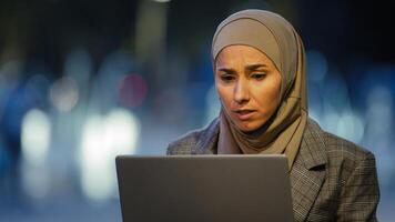 Portrait of muslim business woman user in hijab sits in evening city outdoors working with laptop worry about problem looking at computer wins says yes. Islamic student girl gets offer online winning photo