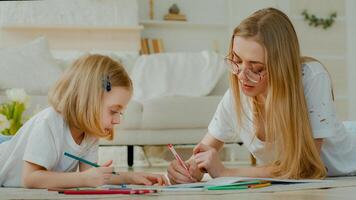 pequeño pequeño adoptado niño niña disfrutar Arte dibujar con caucásico madre en lentes acostado a hogar piso juntos. hija bebé niño con mamá mamá niñera profesor dibujo imagen con lapices hablando foto