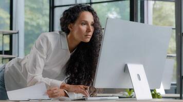 Young attractive hispanic woman professional manager developer working in modern office focused businesswoman checking electronic information on computer using paper documents researching sales data photo