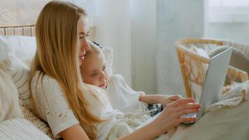 caucásico madre con hija hojeada ordenador portátil en línea Tienda acostado en cama comprando en Internet compras reloj película a dormitorio juntos. mamá enseñar pequeño pequeño bebé niño niña utilizando computadora ciberespacio foto