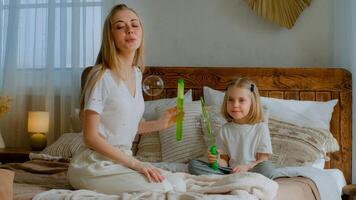 Caucasian mother woman mom with small little child girl kid baby adopted healthy daughter blowing soap bubbles in air in bedroom sitting on bed playing together blow at home family health insurance photo