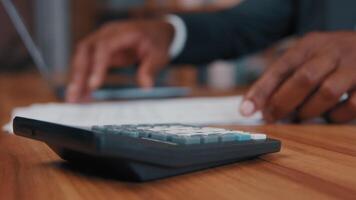 Close-up african american man calculates salary enters data in laptop accountant businessman makes calculation looks for information in paper documents controls business expenses focus on calculator photo