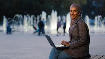 Busy successful muslim student girl in hijab young islamic business woman using laptop app working in city fountains background looking at camera happy smiling. Ethnic female with wireless computer photo