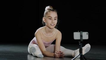 niña adolescente niño bailarina acróbata gimnasta se sienta en piso mirando a cámara web tiene en línea lección danza clase con Internet entrenador acecho en teléfono inteligente en trípode señalando demostración uno índice dedo foto