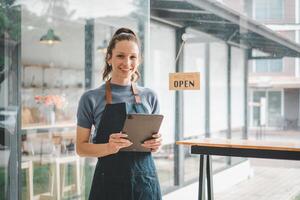 hermosa joven barista mujer en delantal participación tableta y en pie en frente de el puerta de café con abierto firmar tablero. negocio propietario puesta en marcha, SME empresario vendedor negocio concepto. foto