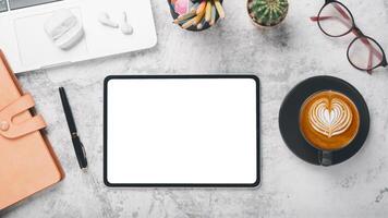 An organized desk featuring a blank tablet screen, elegant coffee cup with latte art, and modern work accessories on a marble surface. photo