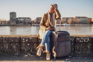 preocupado mujer turista con maleta y teléfono perdido en un ciudad sentado por el río y pensamiento. tonificado imagen. foto