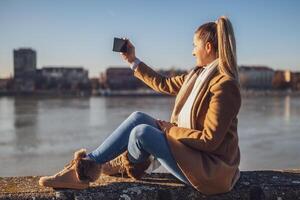 Beautiful woman enjoys photographing with phone cityscape while resting by the river on a sunny winter day. Toned image. photo