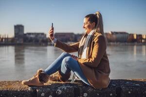 Beautiful woman in warm clothing taking selfie with phone and enjoys resting by the river on a sunny winter day.Toned image. photo