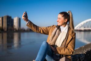 hermosa mujer en calentar ropa tomando selfie con teléfono y disfruta descansando por el río en un soleado invierno día. foto