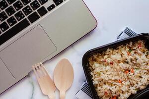 delicioso frito arroz en almuerzo caja en mantelería con de madera cuchara y tenedor y computadora ordenador portátil a lugar de trabajo foto