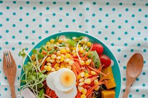 Fresh salad vegetable with boiled chicken egg, wooden spoon and fork on blue polka dot fabric or tablecloth background photo