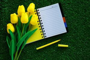 Blank notebook with artificial yellow tulips and a pen on green grass background photo