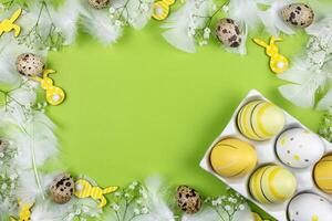 Easter border flat lay with colorful eggs in a white ceramic holder, spring flowers, quail eggs, yellow bunnies and white feathers on a green background. Top view. Copy space. photo