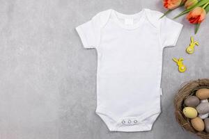 White baby girl or boy bodysuit mockup flat lay with orange tulips flowers and easter eggs decoration on the gray concrete background. Design onesie template, print presentation mock up. Top view. photo