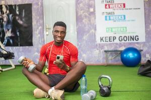 handsome african man in the holding dumbbell smiling as he uses his phone to watch tutorials photo