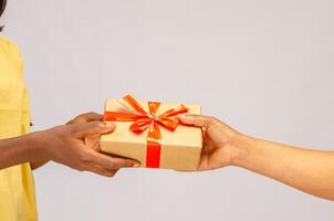 Holiday Celebration Concept. Closeup on female hands holding gift with red bow and giving present box to african american woman, greeting with Birthday, Christmas and New year at home photo
