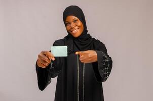 muslim woman holding credit card and smiling at camera isolated on grey photo