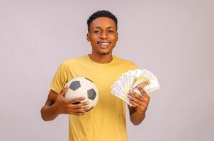 deporte apuestas retrato de emocionado negro chico con fútbol americano pelota y dinero en pie terminado beige fondo, alegre fútbol aficionados celebrando éxito con naira dinero en efectivo, emocionalmente reaccionando a ganar foto