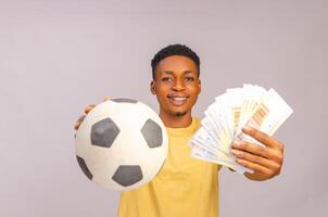 deporte apuesta excitación, apostar y familia estilo de vida concepto. emocionado afroamericano joven chico disfrutando su ganar, en pie con fútbol pelota y un montón de dinero foto