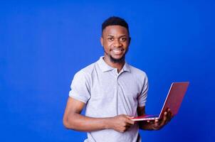 Young smiling african american man standing and using laptop computer isolated over blue background. photo