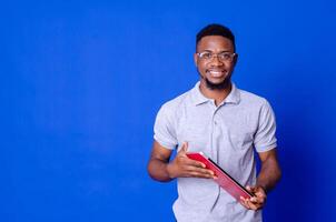 Young smiling african american man standing and using laptop computer isolated over blue background. photo