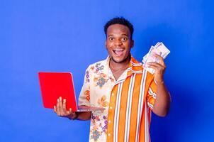 Photo of a happy young afro american handsome man posing isolated over blue wall background using laptop computer holding money.