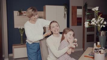 reír, mamá polvos su hija con un cepillo mientras abuela peines su pelo en frente de el espejo. Tres generaciones de mujer. el historia de uno contento familia. video