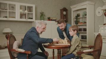 Three generations of men from the same family play chess at home. The grandson makes a move by rearranging a piece on the chessboard. Dad and Grandpa are watching. A family hobby. video