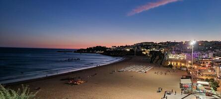 panorámico ver de un costero pueblo a oscuridad con iluminado calles, un sereno playa, y un tranquilo mar debajo un degradado cielo. foto