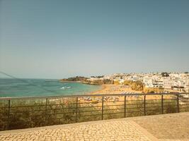 Coastal view of a Mediterranean town with clear blue waters, white buildings, and a sandy beach, seen from a balcony with railing. photo