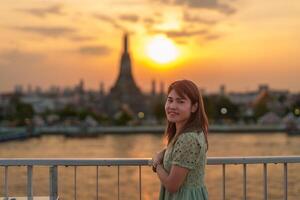 tourist woman enjoys view to Wat Arun Temple in sunset, Traveler visits Temple of Dawn near Chao Phraya river from rooftop bar. Landmark and Travel destination in Bangkok, Thailand and Southeast Asia photo