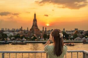 tourist woman enjoys view to Wat Arun Temple in sunset, Traveler take photo to Temple of Dawn by smartphone from rooftop bar. Landmark and Travel destination in Bangkok, Thailand and Southeast Asia