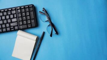 workspace desk with notebooks, pens, keyboard, and glasses on blue background photo