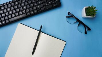 workspace desk with notebooks, keyboard, pens, glasses, plants on blue background photo