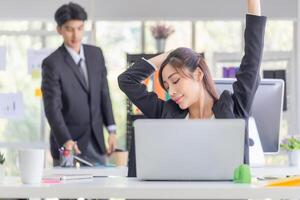 asiático mujer haciendo tramo ejercicio extensión su brazos en trabajo tiempo, hembra oficial sensación cansado y fatiga extensión a relajarse con borroso colegas en moderno oficina foto