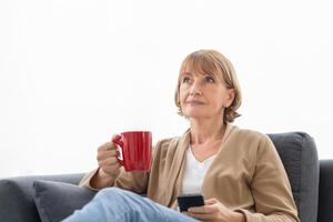 Portrait of Relaxing woman with cups of coffee on cozy sofa in the living room, Mature woman using mobile smartphone during a coffee break photo