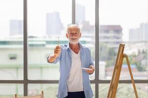 Cheerful senior man showing paintbrush and painting on canvas at home, Elderly man painting on a canvas, Happy retirement concepts photo