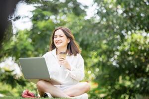 Female freelancer with a laptop and takeaway coffee, headphones sitting in a green park. Smiling young woman looking at a camera and using a laptop computer in the park photo